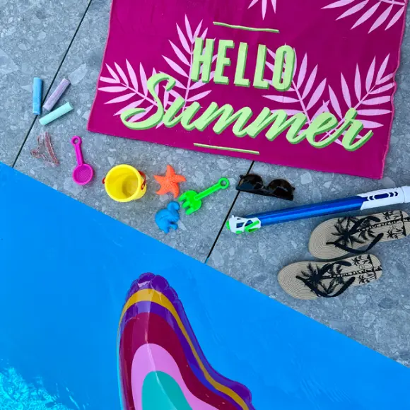 Eine lebhafte Sommerszene mit einem rosa Handtuch mit der Aufschrift „Hello Summer“, Strandspielzeug, Sonnenbrillen und Flip-Flops neben einem Pool.