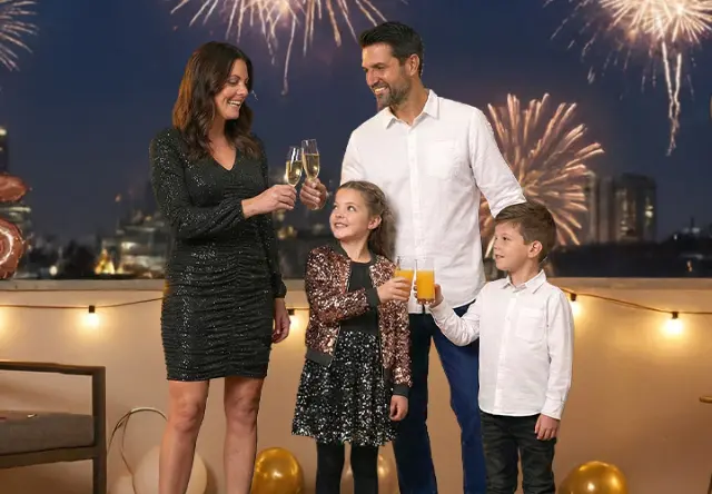 Eine Familie feiert Silvester auf einem Balkon, der mit Luftballons und Lichtern geschmückt ist, während Feuerwerke den Nachthimmel erleuchten.
