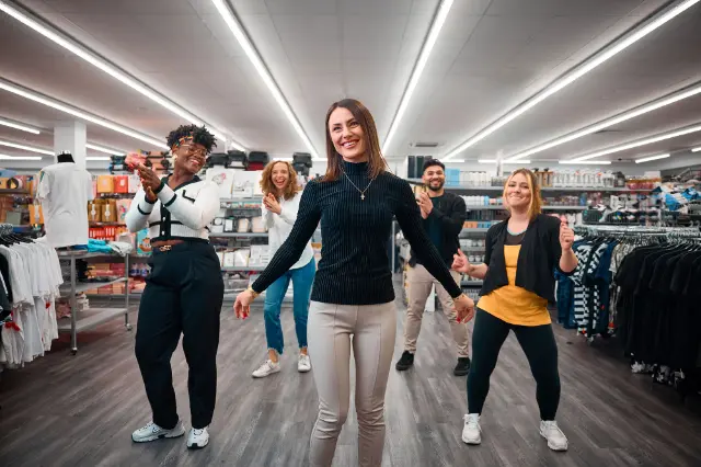 Gruppe von Personen tanzt und klatscht in einem Bekleidungsgeschäft, umgeben von Regalen mit Kleidung.