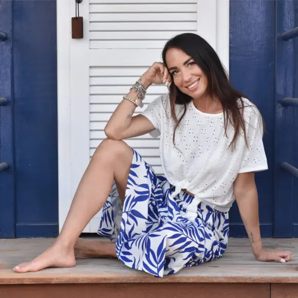 Eine Frau sitzt anmutig auf einer Veranda, trägt ein weißes Oberteil und einen blauen, geblümten Rock, die Hand auf dem Kinn, tief in Gedanken versunken.