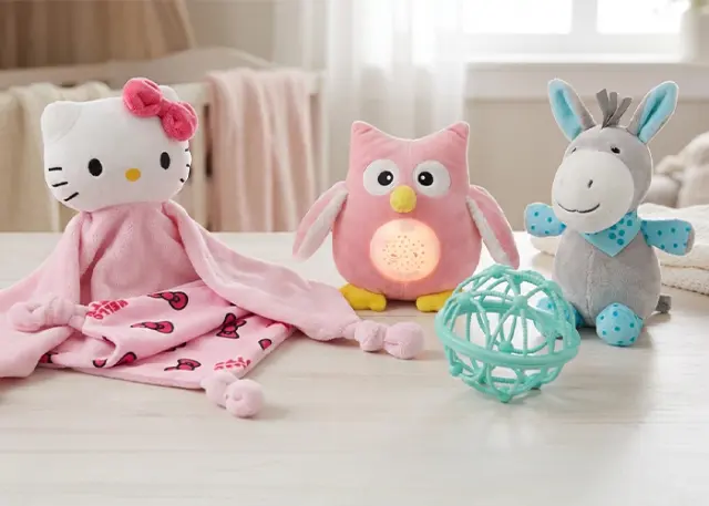 Eine gemütliche Anordnung von Plüschtieren: eine Hello-Kitty-Decke, eine rosa Eule mit Licht, ein grauer Esel und ein petrolfarbener Ball, alles auf einem Holztisch.