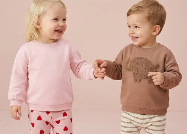 Zwei kleine Kinder halten sich an den Händen, eines trägt einen rosa Pullover und gestreifte Hosen mit Herzmuster, das andere einen braunen Pullover mit Elefantenmotiv.