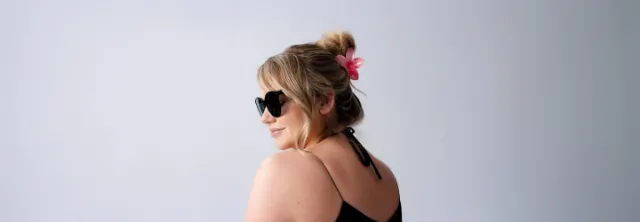 Eine Frau mit gewelltem Haar, das zu einem mit einer rosa Blume geschmückten Dutt frisiert ist und ein schwarzes Oberteil trägt, posiert von hinten vor einem hellen Hintergrund.