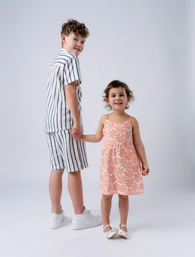 Ein Junge in einem gestreiften Outfit und ein Mädchen in einem geblümten Kleid halten sich lächelnd an den Händen, vor einem schlichten weißen Hintergrund.