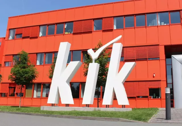 Modernes Gebäude mit leuchtend roter Fassade mit dem großen weißen „kik“-Logo und Eingang, umgeben von Grün an einem sonnigen Tag