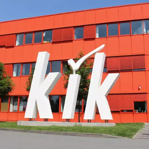 Modernes Gebäude mit leuchtend roter Fassade mit dem großen weißen „kik“-Logo und Eingang, umgeben von Grün an einem sonnigen Tag
