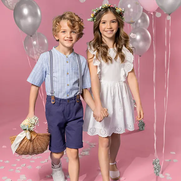 Zwei Kinder halten sich an den Händen und stehen vor einem rosa Hintergrund. Das eine trägt ein gestreiftes Hemd und Shorts, das andere ein weißes Kleid und eine geblümte Krone.