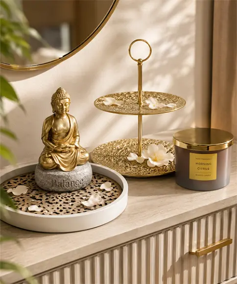 Goldene Buddha‑Figur auf einem „Balance“-Stein, daneben ein goldenes Stufentablett mit Blütenblättern sowie eine Duftkerze „Morning Citrus“ auf einem Holzschrank.