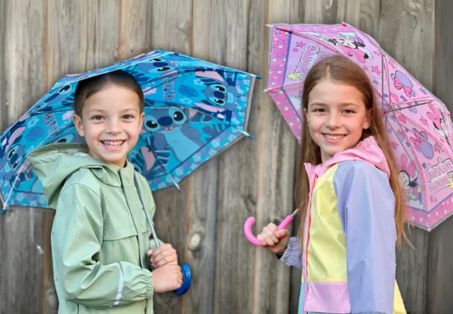 Zwei Kinder stehen lächelnd unter Regenschirmen vor einem Holzzaun. Das Mädchen hält einen rosa Minnie-Mouse-Regenschirm, der Junge einen blauen Stitch-Regenschirm. Sie tragen bunte Regenjacken.