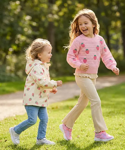 Zwei Kinder laufen fröhlich durch einen sonnendurchfluteten Park, umgeben von grünem Gras und Bäumen, und tragen bunte Pullover und Freizeithosen.