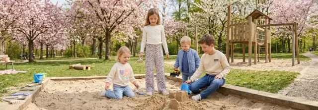 Vier Kinder spielen und bauen Sandburgen in einem Sandkasten unter blühenden Bäumen in einem Park.