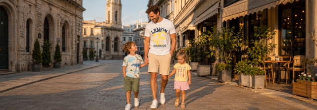 Ein Mann in einem Zitronen‑Print‑Shirt läuft Hand in Hand mit einem Jungen im Dinosaurier‑Print und einem Mädchen im Eiscreme‑Print auf einer Stadtstraße.