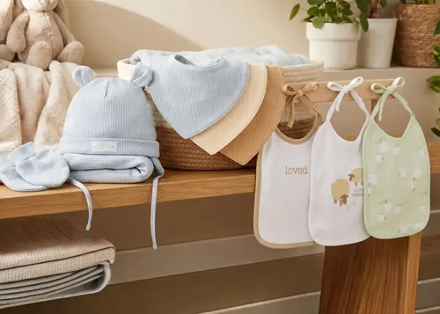 Eine gemütliche Präsentation von Baby-Essentials: eine blaue Mütze, weiche Decken und Lätzchen mit niedlichen Motiven, arrangiert auf einem Holzregal.