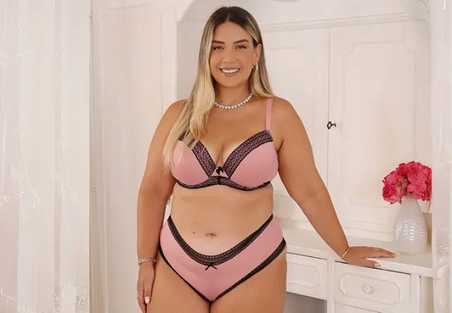 Eine Frau steht selbstbewusst in einem rosa Dessous-Set mit schwarzen Akzenten in einem sanft beleuchteten Raum und trägt eine Perlenkette als Accessoire.