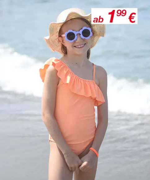 Ein Kind im korallfarbenen One-Shoulder-Badeanzug mit Rüschen steht am Strand und trägt einen breitkrempigen Strohhut. Ein Preisschild ist sichtbar.