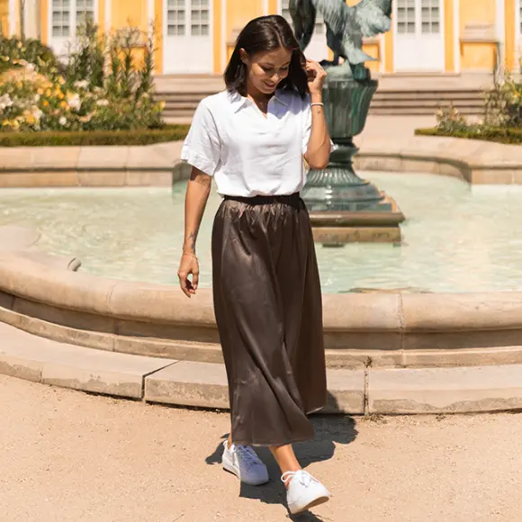 Eine Frau in weißem Shirt und braunem Rock geht an einem Brunnen entlang, umgeben von Grünflächen, mit gelben Gebäuden im Hintergrund.