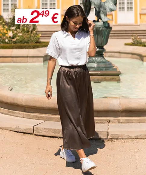 Eine Frau in weißem Shirt und braunem Rock geht an einem Brunnen entlang, umgeben von Grünflächen, mit gelben Gebäuden im Hintergrund.