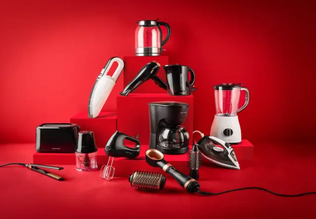 Eine Sammlung verschiedener Haushaltsgeräte, darunter ein Wasserkocher, ein Mixer, eine Kaffeemaschine und ein Staubsauger, arrangiert auf roten Stufen.