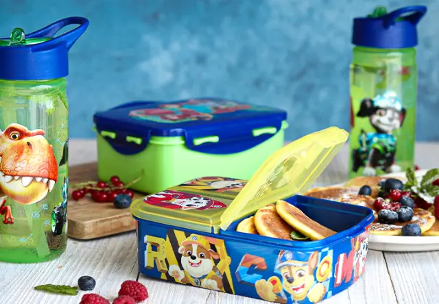 Brotdose und Trinkflaschen mit PAW Patrol und Dinosauriermotiven auf einem gedeckten Tisch mit Snacks und Obst für Schule oder Kindergarten. Brotdose und Trinkflaschen mit PAW Patrol und Dinosauriermotiven auf einem gedeckten Tisch mit Snacks und Obst für Schule oder Kindergarten.