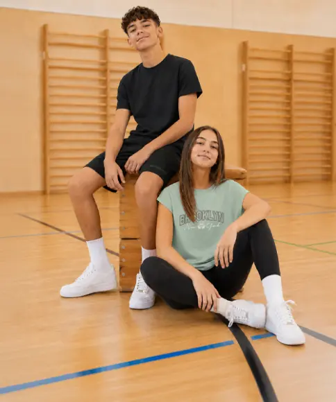 Zwei Jugendliche tragen bequeme Sportbekleidung und sitzen in einer Turnhalle auf und neben einem Sprungkasten. Die Outfits eignen sich ideal für den Schulsport.