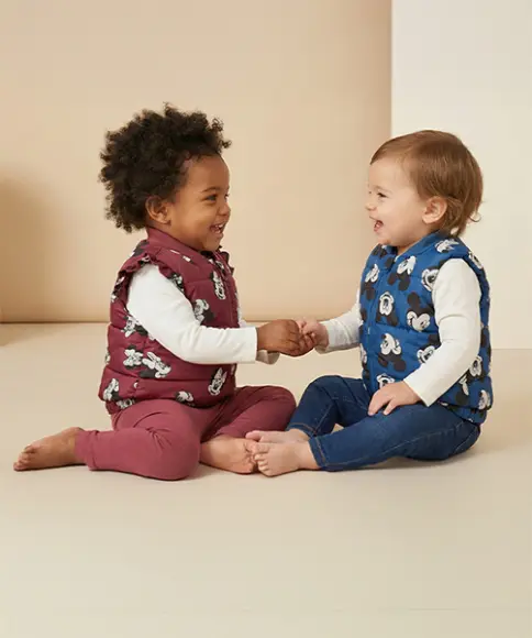 Zwei Kleinkinder sitzen vor einem hellen Hintergrund, tragen bunte Westen, halten sich an den Händen und lächeln sich gegenseitig an.
