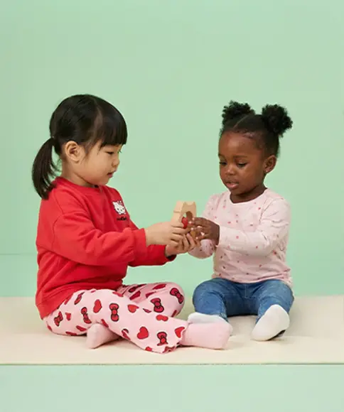Zwei kleine Kinder sitzen auf einer weichen Unterlage und spielen fröhlich miteinander, während sie ein buntes Spielzeug vor einem pastellgrünen Hintergrund halten.