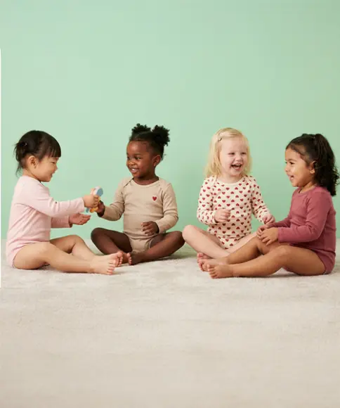 Eine Gruppe von vier Kleinkindern sitzt auf einem hellen Boden und beschäftigt sich gemeinsam mit einem Spielzeug vor einem einfarbigen pastellgrünen Hintergrund.