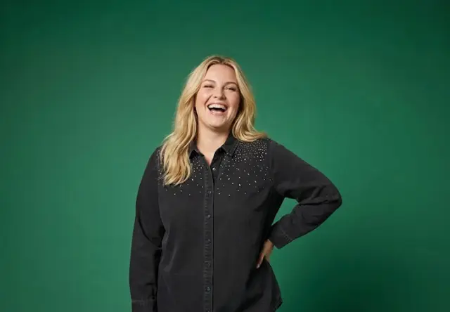 Eine Frau mit welligem blondem Haar steht selbstbewusst in einem schwarzen Shirt mit dezenten Verzierungen vor einem grünen Hintergrund.