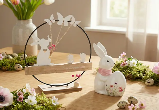 Eine dekorative Tischdekoration mit einer weißen Hasenfigur, einem Holzdisplay mit Schmetterlingen und floralen Akzenten auf Grünpflanzen. Eine dekorative Tischdekoration mit einer weißen Hasenfigur, einem Holzdisplay mit Schmetterlingen und floralen Akzenten auf Grünpflanzen.