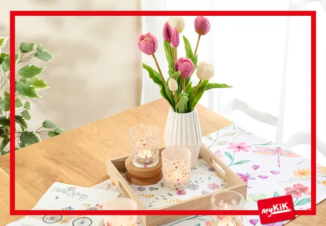 Ein Holztisch mit rosa und weißen Tulpen in einer Vase, umgeben von Kerzen und Blumenschmuck. Ein blumiges Tischset und ein Zettel sind ebenfalls zu sehen.