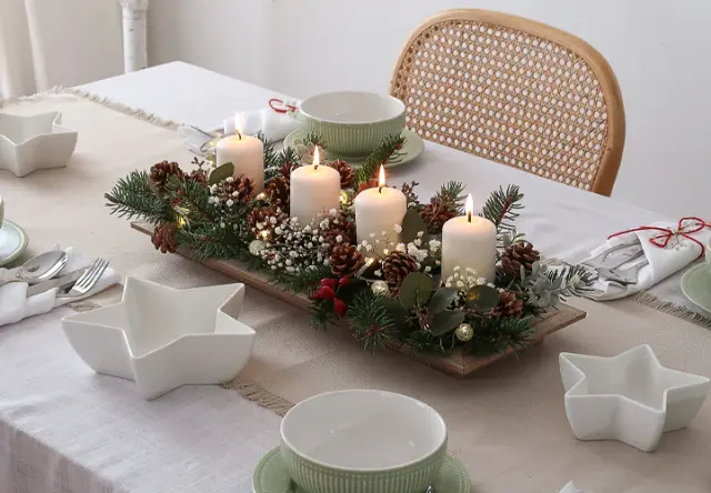 Ein festlich gedeckter Tisch mit einem Mittelstück aus brennenden Kerzen, umgeben von Grün, Tannenzapfen und weißen Blumen auf einer neutralen Tischdecke.