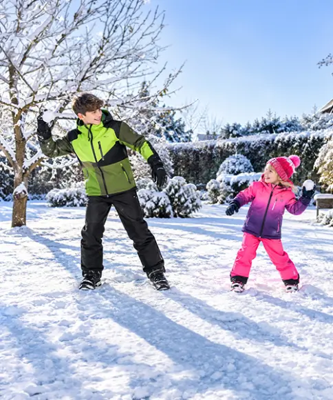 Junge in grün-schwarzer Skijacke und Mädchen in pinker Skibekleidung spielen mit Schneebällen im verschneiten Garten