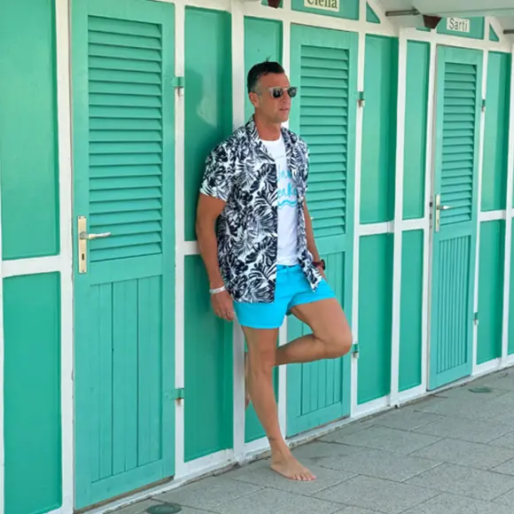 Ein Mann in einem tropischen Hemd und blauen Shorts lehnt lässig an grünen Strandhütten und genießt den sonnigen Tag.
