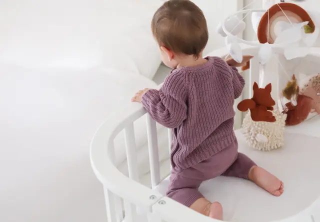 Ein Kleinkind in einem gemütlichen lila Outfit steht in einem weißen Kinderbett und betrachtet ein Mobile mit verspielten Tierfiguren.