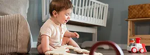 Sitzendes Baby spielt mit Holzspielzeug in gemütlichem Kinderzimmer mit Bettchen und Spielauto.