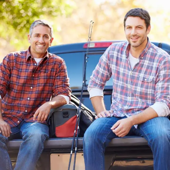 Zwei Männer sitzen auf der Heckklappe eines Lastwagens und genießen einen sonnigen Tag im Freien, umgeben von Bäumen, mit Angelausrüstung in der Nähe.