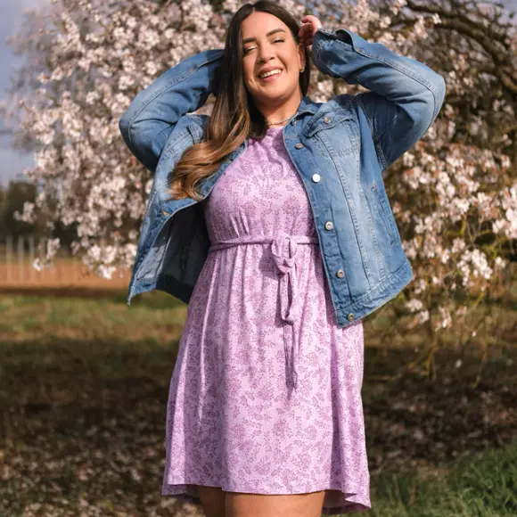 Eine Frau in einem lila Kleid und einer Jeansjacke posiert vor einem blühenden Baum.