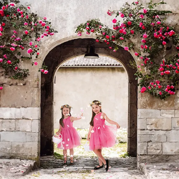 Zwei Mädchen in rosa Kleidern stehen unter einem Blumenbogen, umgeben von leuchtend rosa Blumen.