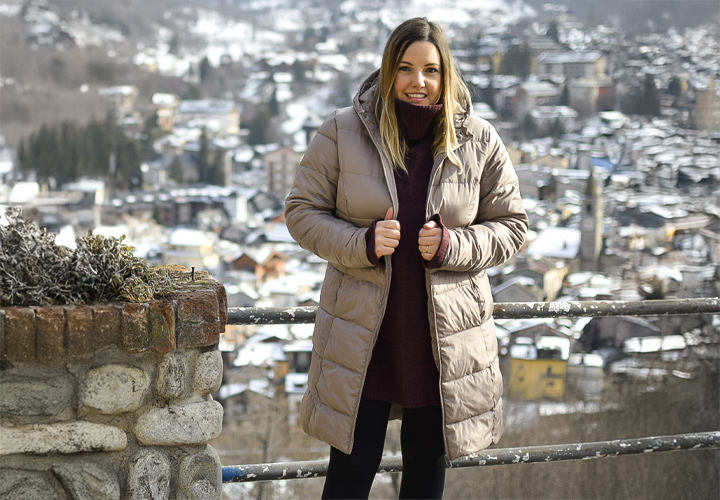 Eine Frau in einem stylischen beigen Daunenmantel steht an einem Steingeländer und blickt auf ein verschneites, malerisches Bergdorf.
