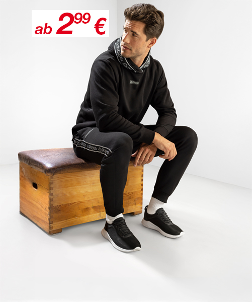 Ein junger Mann in einem schwarzen Kapuzenpulli und Jogginghose sitzt auf einer Holzbank, trägt Sportschuhe und hat ein Preisschild über sich angebracht.