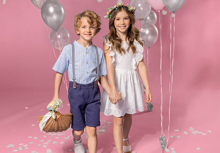 Zwei Kinder halten sich an den Händen und stehen vor einem rosa Hintergrund. Das eine trägt ein gestreiftes Hemd und Shorts, das andere ein weißes Kleid und eine geblümte Krone.