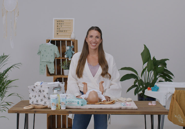 Eine Frau steht an einem Holztisch mit Babypflegeprodukten, Kleidung und Spielzeug, umgeben von Grünpflanzen in einer hellen Umgebung. Eine Frau steht an einem Holztisch mit Babypflegeprodukten, Kleidung und Spielzeug, umgeben von Grünpflanzen in einer hellen Umgebung.