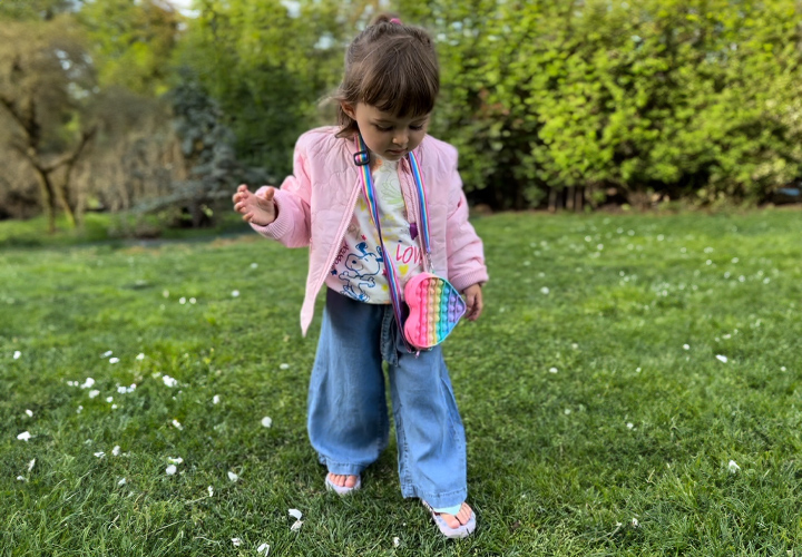 Ein Kind in einer rosafarbenen Jacke und einer bunten Tasche steht selbstbewusst auf einem grasbewachsenen Boden