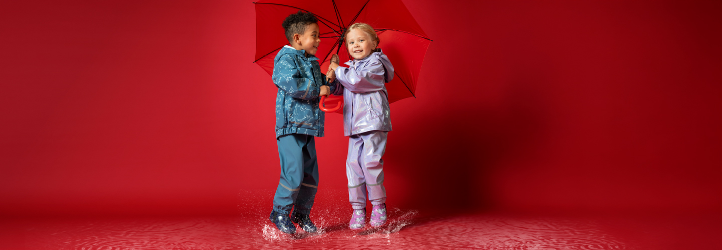 Zwei Kinder in bunten Regenmänteln planschen fröhlich in Pfützen, während sie sich einen leuchtend roten Regenschirm teilen.