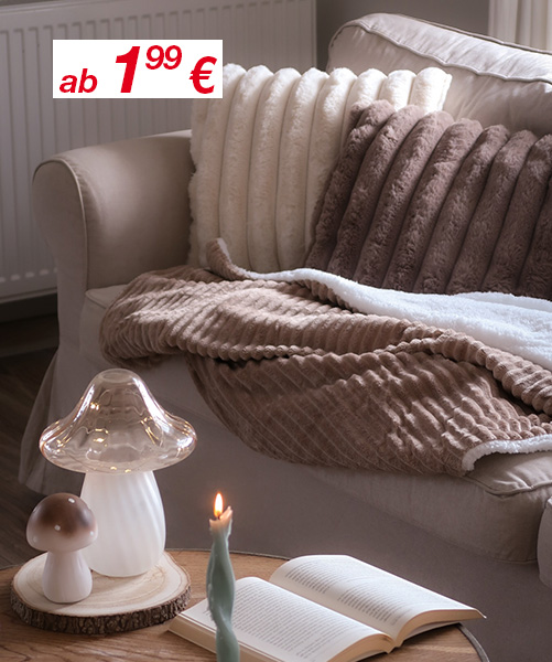 Gemütliche Wohnzimmerszene mit weichen Kissen, flauschigen Decken, einer leuchtenden Kerze und einer Pilzlampe auf einem Holztisch.
