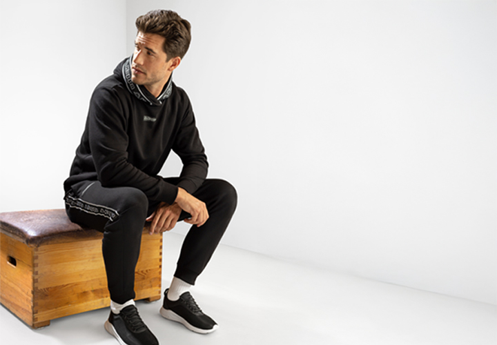 Ein Model steht in einem eng anliegenden schwarzen Hoodie und einer passenden Jogginghose vor einem leuchtend roten Hintergrund, die Hände in den Taschen, und trägt sportliche Sneakers.