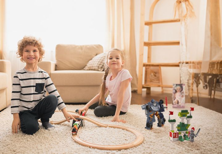 Zwei Kinder spielen auf einem Teppichboden mit einer Holzeisenbahn, einem Spielzeugroboter und einem Baukasten, umgeben von einem gemütlichen Wohnzimmer.