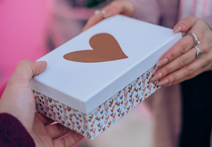 Eine Person überreicht eine dekorative Geschenkschachtel, die mit einem Herz und bunten Mustern verziert ist