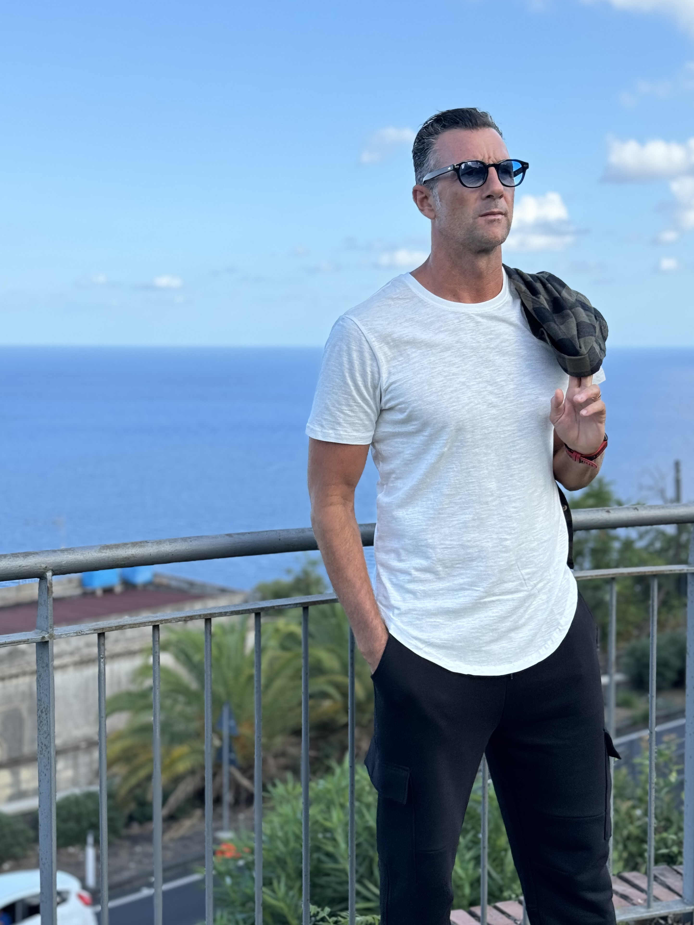 Mann mit weißem T-Shirt und schwarzer Hose, trägt Sonnebrille und Jacke über der Schulter