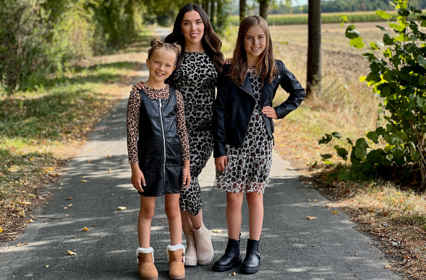 Leoparden Muster Drei Kinder posieren auf einem mit Blättern übersäten Weg, gekleidet in stilvolle Outfits mit Leopardenmuster, umgeben von Bäumen und einem klaren Himmel über ihnen.